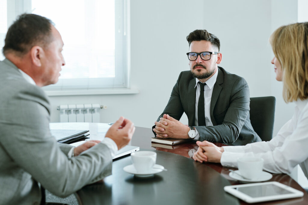 Mature serious businessman in formalwear and his young colleague sitting in front of director of company and listening to what he saying